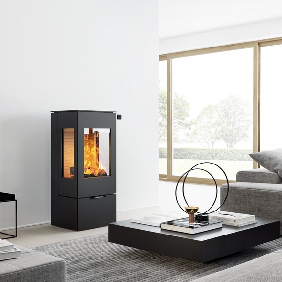 Holz-Kaminofen NEXO 100 in Schwarz mit Stahltür und zwei Seitenfenstern in einem modernen, hellen Wohnraum