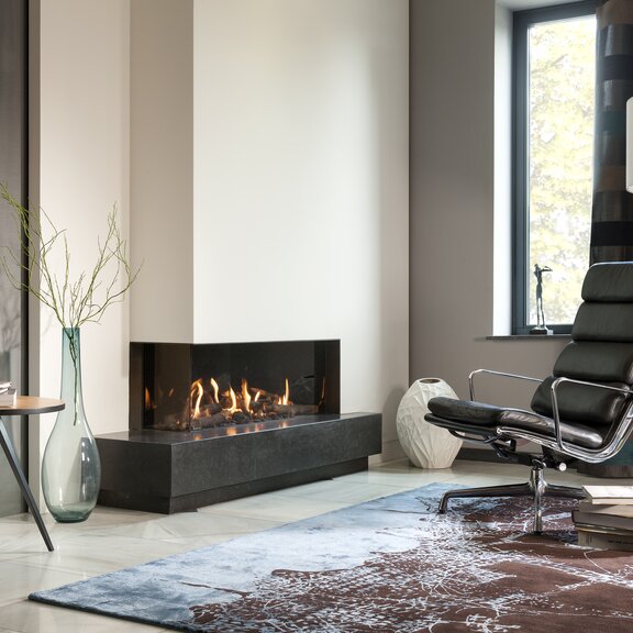 Gas-Kamin G105/37C Eckkamin links von Kalfire mit Keramik Holzscheiten im Feuerraum in einer modernen Wohnung mit Sessel in Leseecke