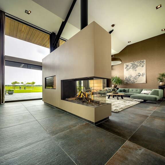 Raumteiler Gas-Kamin GP115/75R mit sandfarbiger Verkleidung im Wohnzimmer mit grossen Fensterfronten und Aussicht in die grüne Landschaft